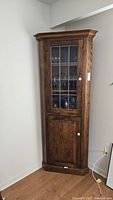 Tall oak corner cabinet placed in corner against walls with visible wood grain on doors and glass display upper door fitted with multiple glass panels.