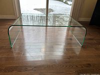 Front view of clear glass coffee table showing top surface and waterfall sides on hardwood floor.