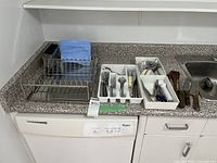 Photo showing kitchen countertop with metal dish rack and three white plastic trays holding various cutlery and kitchen utensils including forks, spoons, knives, scissors, tongs, and wooden spoons.