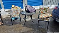 Full patio set including table with glass top, four teal-colored chairs, floral cushions, umbrella stand base, and cover