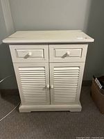 Front view of the off white antiqued wooden nightstand with 2 drawers and 2 louvered doors beneath, placed against a wall on carpeted floor, showing its condition and shape.