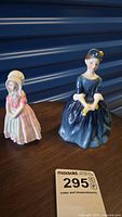 Two Royal Doulton figurines on dark wooden surface, Tootles in pink dress on left and Cherie in blue dress on right.