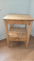 Front view of older wooden side table showing rectangular top, single drawer with knob, and lower shelf made of wooden slats on wooden legs, with visible wear and aging.