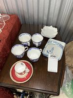 Collection of 4 blue and white Royal Crown Derby teacups, 1 red and gold Manoir Richelieu teacup with saucer, 2 white plates, and a blue and white handled dish on wooden table.