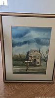 Full view of framed watercolour farmhouse painting with sky and bare trees.