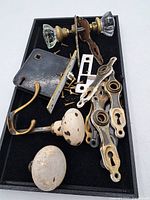 Tray with various antique door hardware including door knobs, backplates, keyhole covers, a metal hook, latch plate, and screws, shown on a black tray.
