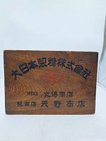 Front side of antique Japanese wooden sugar crate cover with black and red Japanese lettering on wood grain background.