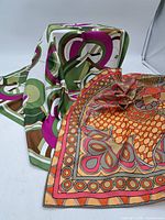 Two silk scarves displayed, one with green and purple geometric pattern and the other with orange and pink floral pattern.