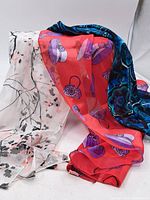 Three scarves laid out showing colors and patterns including white with black and pink print, red with purple handbag and hat motifs, and blue floral design.