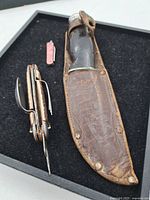 Photo showing three vintage utility cutters including fixed blade knife with black handle in leather sheath, folding multi-tool cutter with knife, fork, and spoon, and small pink folding knife.