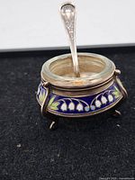Photo showing vintage open salt cell with blue and white floral enamel design, seated on three legs, with gold-tipped spoon placed inside.