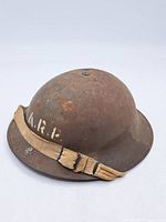 Front and side view of the rusty steel ARP helmet showing beige cloth chin strap and front 'A.R.P.' painted marking.