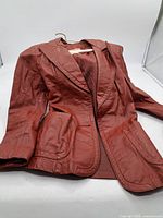 Photo of maroon women's leather jacket laid flat showing front, two front pockets, lapel collar, and fabric lining.
