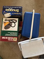 Multiple items in the lot: Snuggie box, Bellows Air Pump box, blue ice cooler with white handle and lid, and a partial view of a white plastic chest or cooler lid.