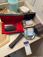 Two vintage straight razors with black cases, black plastic shaver holder, electric shaver in red velvet case, bathroom items including small round mirror and absorbent cardboard pack, along with an electric shaver charger in blue case.