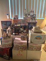 Photo showing multiple ceramic miniature cottages stacked on and around their original branded David Winter boxes, arranged on a carpeted floor near a vertical blind window and table lamp.