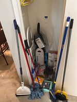 Cleaning supplies including brooms, mop, dustpan, upright and handheld Hoover vacuums in closet corner