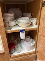 Overview of cabinet shelf displaying stacked Centura dishware including plates, bowls, cups, and martini glass. Bottom shelf shows utensil carousel and glass jars with lids.