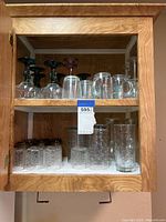 Photo showing top and bottom shelves in a cabinet with assorted drinking glasses including clear drinking glasses and colored-stem goblets and wine glasses.