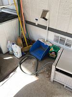 Wide view of gardening tools neatly lined against wall including brooms, rake, blue hose holder, hose, and various bottles.