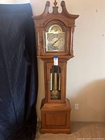 Full view of the grandfather clock showing wooden case with decorative top and base, sitting against a wall.