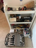 Photo showing white plastic three-tier shelf organizer holding various tools including hammers, scissors, broom, screwdrivers, flashlight, garden tools, and small hardware accessories with additional items on floor beside organizer.