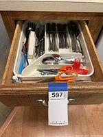 Photo showing the kitchen drawer containing stainless steel forks, knives, spoons, an orange-handled scissors, blue pliers, and a manual can opener.