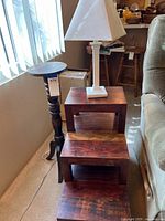 Photo showing a vintage wooden lamp stand with turned legs, a small white table lamp with a square base and white shade, and a dark wooden stairstep end table with three steps.
