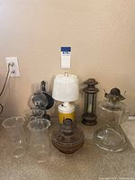 Full view of various lamps arranged against a wall showing multiple oil lamps, battery lamp, and glass chimneys.