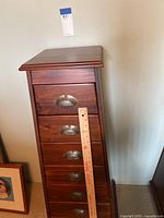 Front view of tall wood lingerie dresser, showing seven drawers and metal cup pulls with fan motif. A wooden ruler is placed vertically in front for scale.