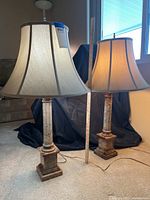 Pair of marble lamps standing on floor near window with measuring stick showing height around three feet. Beige bell-shaped fabric shades with dark edges. Marble bases and columns with mottled coloration.