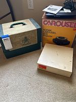 Vintage Brumberger slide projector carrying case, Kodak Carousel 600 projector box and a beige box, all placed on carpet.