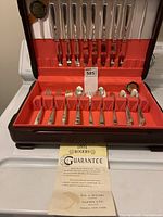 Full view of the opened wooden case showing the flatware neatly arranged in slots, red felt interior, and a guarantee paper on display.