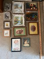 Collection of 13 framed artworks laid out on carpet with a yardstick showing size; includes landscapes, animals, people, and nature scenes in varying frame styles and sizes.