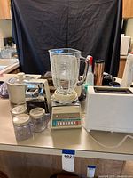 Wide photo showing the small kitchen appliances grouped on a kitchen counter including blender, two toasters, crockpot, electric casserole, can opener and drinkmaster coffee maker