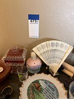 Photo of assorted small decorative items on dark tabletop including cranberry glass covered dish, ceramic jar, white folding fan, and framed artwork attached to wall