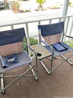 Two folding camping chairs outside showing navy and beige fabric and metal frames with armrests and cup holders.