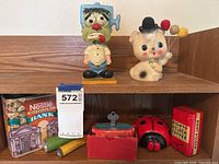 Shelf display of six assorted vintage coin banks including Nestle Hot Cocoa Mix, ladybug, clown, coffee tin, Frankenstein figurine, and green cylindrical bank.