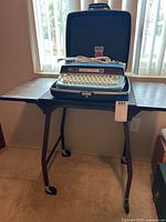 Blue Coronet electric typewriter in a matching blue hard case set on the foldable rolling cart with four caster wheels.