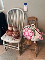 White painted wooden chair with worn and peeling paint, holding two brown pottery vases with geometric patterns and a large white piggy bank ceramic with pink flowers, next to a brown wooden chair not included.