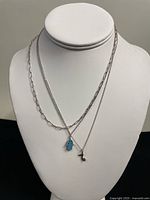 Two silver necklaces displayed on a bust stand showing paperclip chain with musical note pendant and a chain with turquoise hamsa hand pendant.