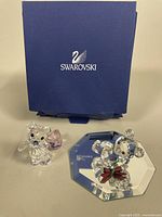 Two Swarovski Kris Bear crystal figurines on beige surface with original blue Swarovski box behind.