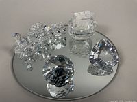 Four Swarovski crystal pieces on a round mirror: crystal train, heart-shaped ring box with frosted flower, faceted crystal heart, and crystal sphere.