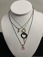 Three silver necklaces on display bust with different pendants. Key, circular rings with charm, and pink gemstone pendants visible.
