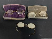 Three pairs of wire-frame spectacles displayed with two original spectacle cases, showcasing age and style from circa 1890s-1910s.