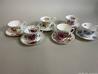 Six assorted bone china teacups and saucers arranged on flat surface showing variety of floral and fruit patterns.