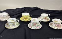 Six floral bone china teacups and saucers displayed on black fabric background, various brands and floral designs.