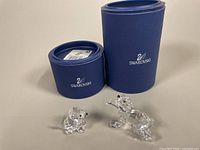 Two Swarovski crystal figurines with their original blue Swarovski cylindrical boxes displayed behind them on a white surface.