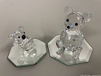 Two Swarovski crystal teddy bear figurines displayed on mirrored octagonal bases. Larger and smaller figurine shown side by side.