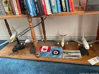 Four airplane models on stands displayed on wooden shelf below books, with small Royal Canadian Air Force flag and black and white photo on the shelf.